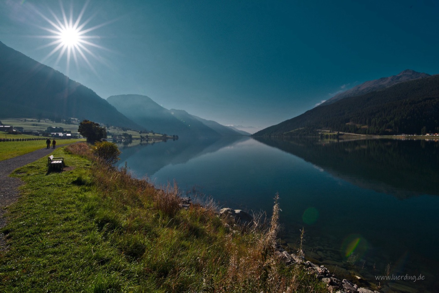 Ruhiger Bergsee mit Spiegelung, die Sonne bricht durch die Bäume am Ufer